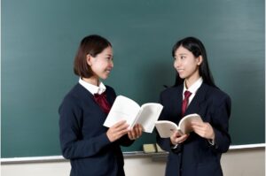 黒板の前でテキストを開きながら会話する2人の女子高生